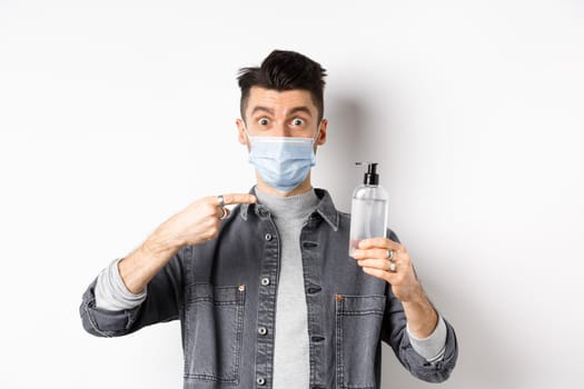 Health, covid and pandemic concept. Stylish guy in medical mask pointing at bottle with hand sanitizer, showing antiseptic, standing on white background.
