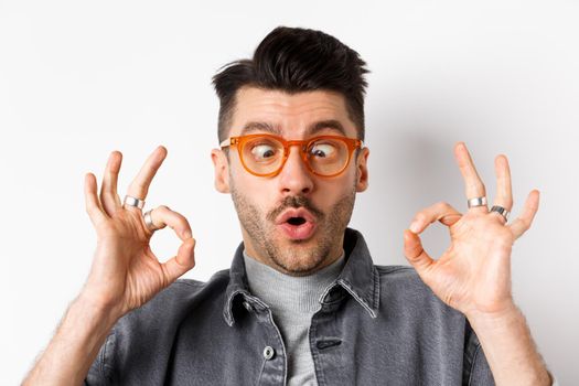 Funny young man in stylish glasses squinting eyes, showing okay gesture, recommend eyewear shop, standing on white background.