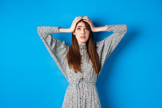 Troubled young woman with long hair, holding hands on head in panic, looking up nervous, having problem, standing on blue background.