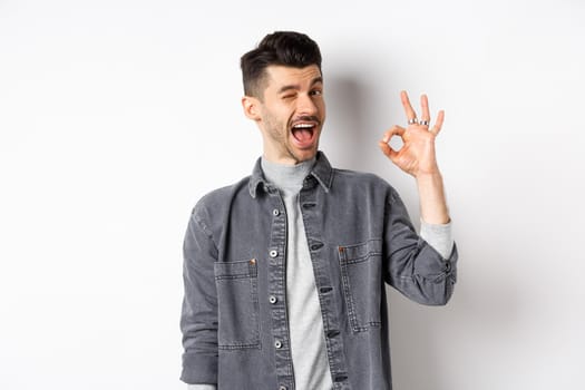 Excited handsome guy with moustache, winking and showing okay sign, smiling pleased, assure all good, praise nice work, making good job gesture, standing against white background.