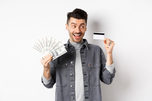Excited guy holding plastic credit card and dollar bills, standing amused on white background.