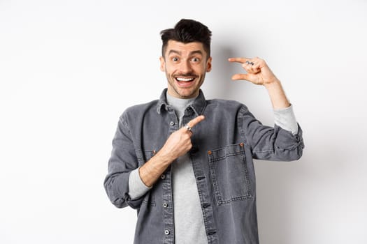 Excited man showing small size, pointing finger at little thing and smiling, standing against white background.