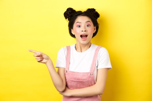 Excited asian teen girl pointing finger left at logo and looking happy, showing advertisement, standing on yellow background.