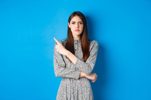 Disappointed frowning girl pointing left and looking displeased, condemn something bad, complaining at banner, standing against blue background in dress.