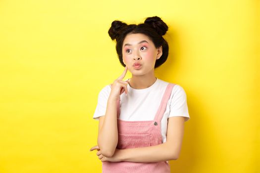 Summer lifestyle concept. Pensive glamour asian girl thinking, looking left at logo and dreaming, making choice, standing on yellow background.