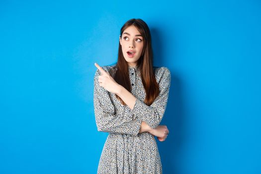 Wow look there. Excited pretty lady in dress looking and pointing left wondered, checking out advertisement, standing on blue background.