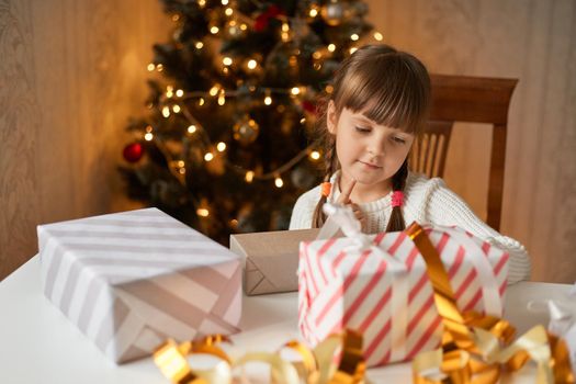 Charming child pack Christmas gifts in festive room, looks at boxes, wearing casual attire, having dark hair an two pigtails, presents for new year tree.