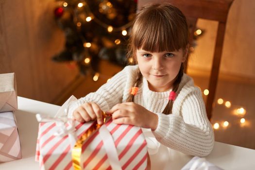 Charming female with braids wearing casual white sweater packing Christmas gifts, looking at camera with cute expression, sitting at table and preparing for new year.