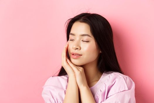 Tender and romantic girl gently touching face, close eyes and smiling softly, standing on pink background. Concept of skincare.