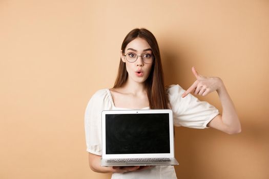 Excited girl in glasses making presentation on computer, pointing hand at laptop screen and say wow, standing on beige background.