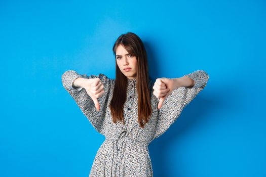 Negative emotion. Disappointed frowning woman with long hair and dress, showing thumbs down and grimacing with dislike, express aversion, standing on blue background.