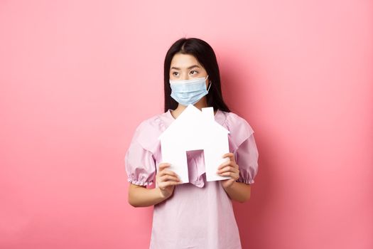 Real estate and pandemic concept. Young asian woman in medical mask dreaming of home, showing paper house cutout and look aside, standing against pink background.