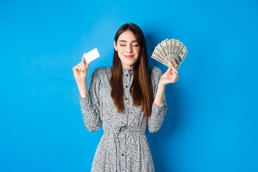 Shopping. Happy smiling woman looking satisfied with eyes closed, showing money dollar bills and plastic credit card, standing dreamy against blue background.