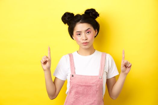 Annoyed and tired asian girl with pink glamour makeup, pointing fingers up and look bothered, standing on yellow background.