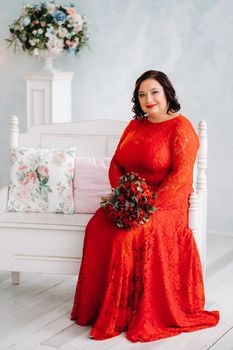 a woman in a red dress sits on a sofa and holds a bouquet of red roses and strawberries in the interior