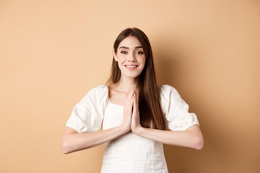 Pretty please. Cute young woman asking for favour, holding hands in begging pose and smiling, need help, standing on beige background.