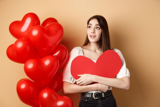 Romantic girl hugging Valentine day big red heart cutout, standing near romance balloons and looking left at logo, gazing dreamy at promo, beige background.