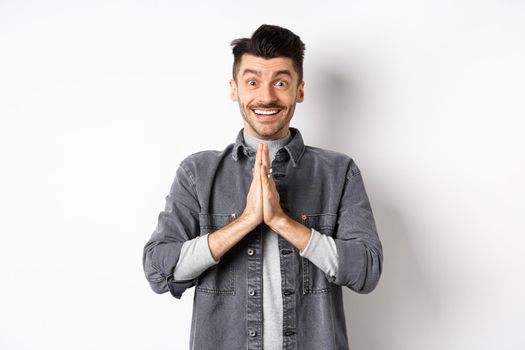 Please help me. Smiling cute man holding hands in namaste gesture, asking for favour, saying thank you, looking hopeful and excited at camera, standing on white background.