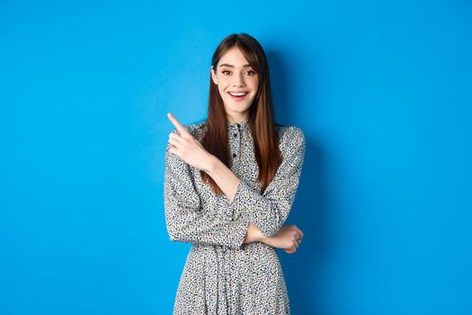 Cheerful young woman in long dress smiling, pointing finger at upper left corner, advertising product on blue background.