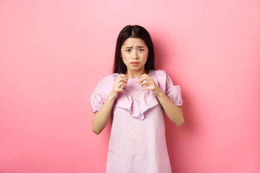 Scared asian woman victim, looking frightened, trembling from fear, stare at scary thing, standing anxious on pink background.