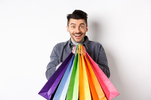 Excited smiling guy holding colorful shopping bags and rejoicing with discounts in store, standing against white background.