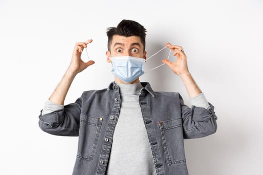 Health, covid and pandemic concept. Funny guy playing with medical mask on face, showing faces, standing against white background.