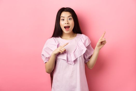 Excited asian teenage girl pointing fingers right, smiling happy and amused, showing cool news, standing on pink background.