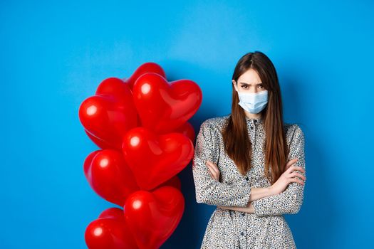 Covid-19, quarantine and health conept. Angry girlfriend in medical mask, standing near Valentines day red heart balloons, cross arms on chest and frowning offended, blue background.
