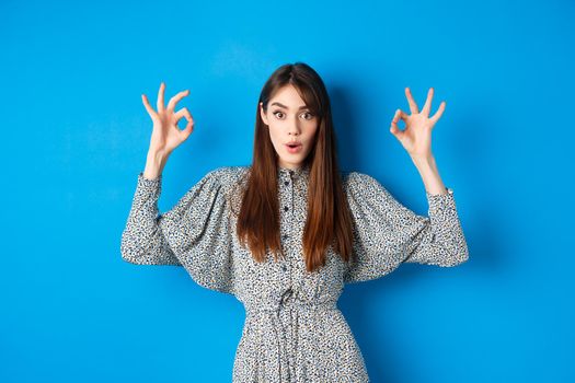 Excited beautiful woman in dress say wow, recommend cool thing, praising and looking impressed, standing on blue background.
