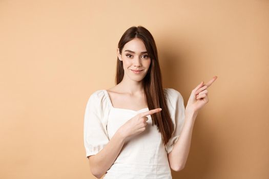 Beautiful young woman in dress pointing fingers right, showing logo and looking tender at camera, beige background.