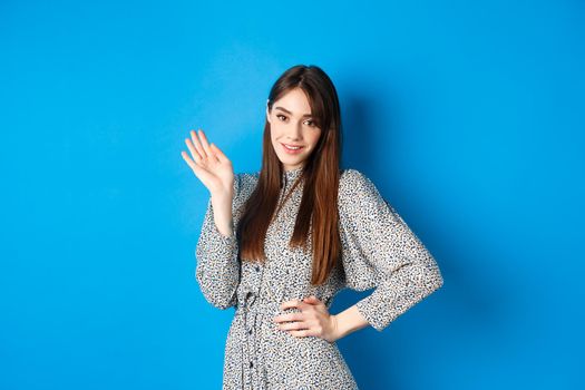 Beautiful lady in dress waving hand and say hello, greeting you with cute smile, standing on blue background.