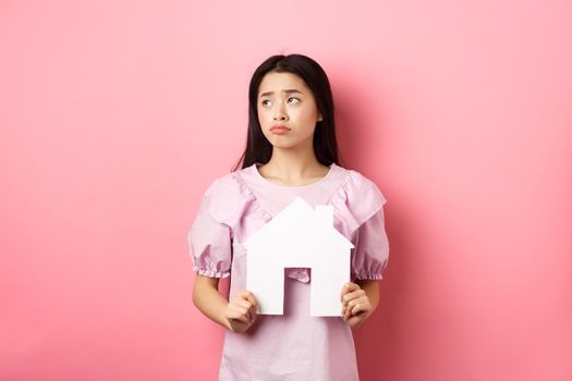 Real estate and insurance concept. Sad asian woman looking aside with unfair face, showing paper house cutout, dreaming of apartment, standing against pink background.