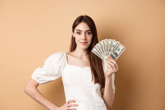 Confident smiling woman showing dollar bills, earn money and look satisfied, standing on beige background with cash.