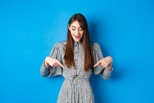 Dreamy happy woman pointing fingers down and looking wondered at promotion, showing awesome news, standing in dress on blue background.