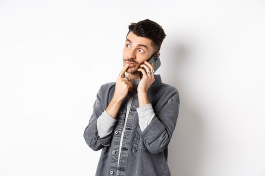 Image of pensive handsome man talking on phone and thinking, making choice while making takeout order, standing against white background.