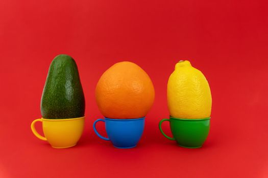 Green avocado at yellow cup, orange at blue cup, yellow lemon at green cup, all of them are on red background. Enough space for text, minimalistic concept. They are different colors, red backdrop.