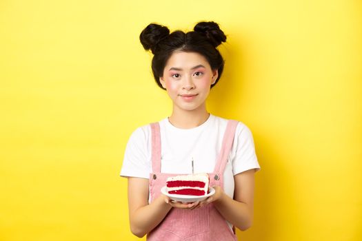 Asian birthday girl standing with cake and smiling, celebrating b-day on yellow background.