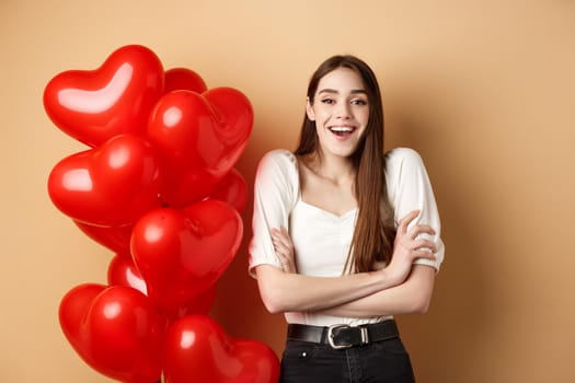 Valentines day concept. Beautiful young woman having fun, laughing and smiling at camera, standing near romantic heart balloons, beige background.