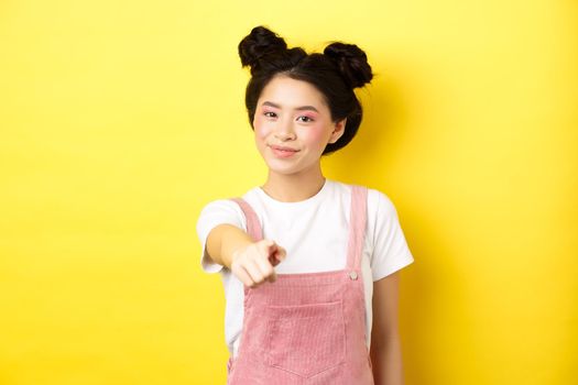 Stylish japanese girl with beauty makeup pointing at camera, smiling at you, beckon and invite people, standing on yellow background.