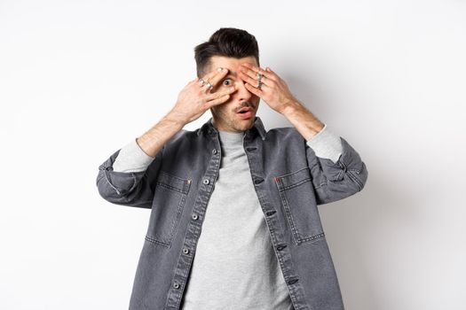 Wow interesting. Curious guy peeking through fingers at camera, looking amazed, standing on white background.