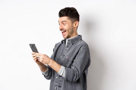 Profile portrait of excited handsome man reading message on phone, looking happy at smartphone screen, standing on white background.