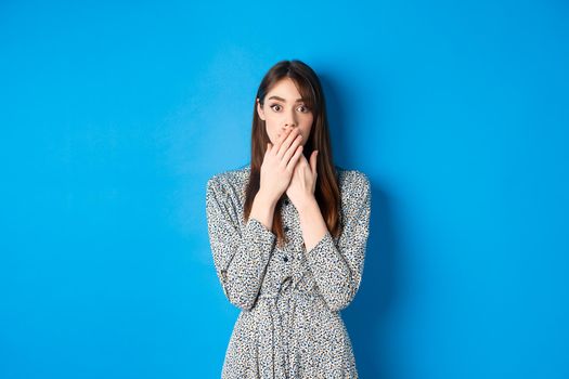 Shocked woman in dress gasping, covering mouth with hands and look excited, hear gossips, standing against blue background.