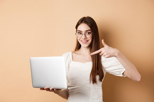Smiling woman in glasses pointing finger at laptop screen, showing online promo, standing on beige background.