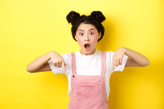 Surprised asian woman with glamour makeup and hairstyle, drop jaw and pointing down amazed, standing against yellow background.