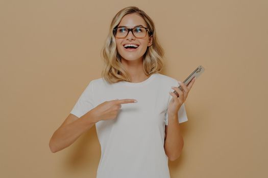 Happy and excited girl in spectacles pointing on mobile phone and looking aside with amazed face expression, reading unbelievable and good news in internet while posing in studio
