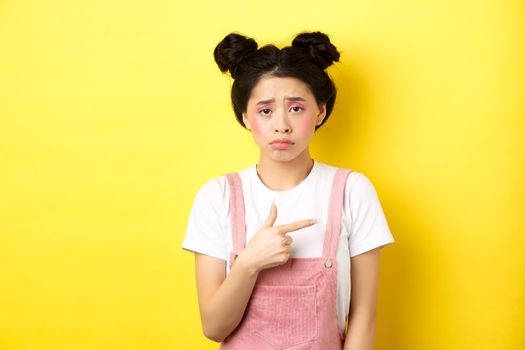 Sad and disappointed asian girl frowning, feel unfair, pointing finger right at bad thing, complaining on something upsetting, yellow background.