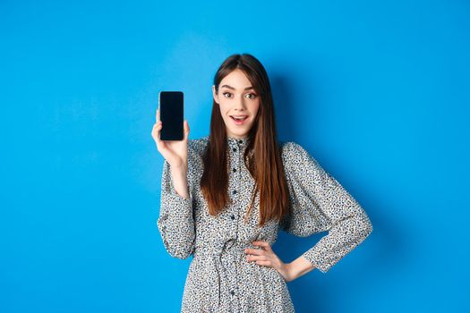Amazed smiling woman showing empty smartphone screen, look impressed, recommending online promo, blue background.