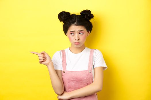 Upset frowning teenage girl sulking, pointing fingers left with jealous unfair face, standing disappointed on yellow background.