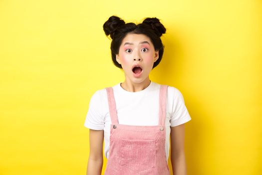 Excited asian teen girl drop jaw, gasping amazed with opened mouth, looking at camera impressed, checking out summer promo offer, standing on yellow background.