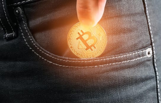 Male hand holding gold bitcoin and putting into pocket of jeans, close-up.